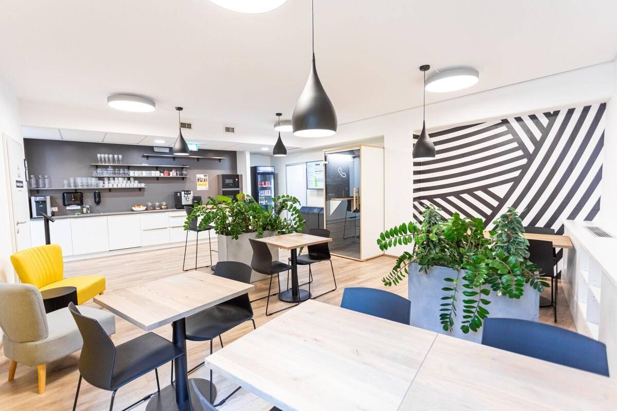 Moderne Büroküche und Essbereich mit Kaffeemaschine, Tischen, Stühlen und vielen Pflanzen.