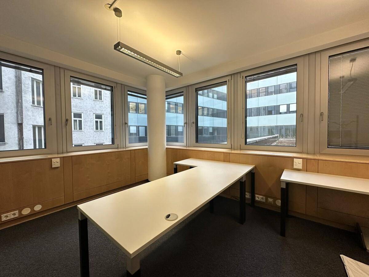 Helles Bürozimmer mit mehreren Fenstern, einem zentralen Pfeiler und modernen Schreibtischen, ideal für konzentriertes Arbeiten.