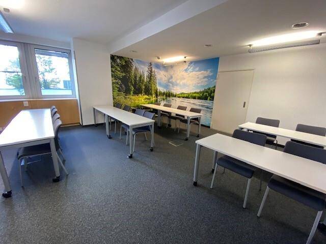 Heller Schulungsraum mit mehreren Tischen und Stühlen, belebt durch eine farbenfrohe Wandtapete mit Wald- und Seemotiv.