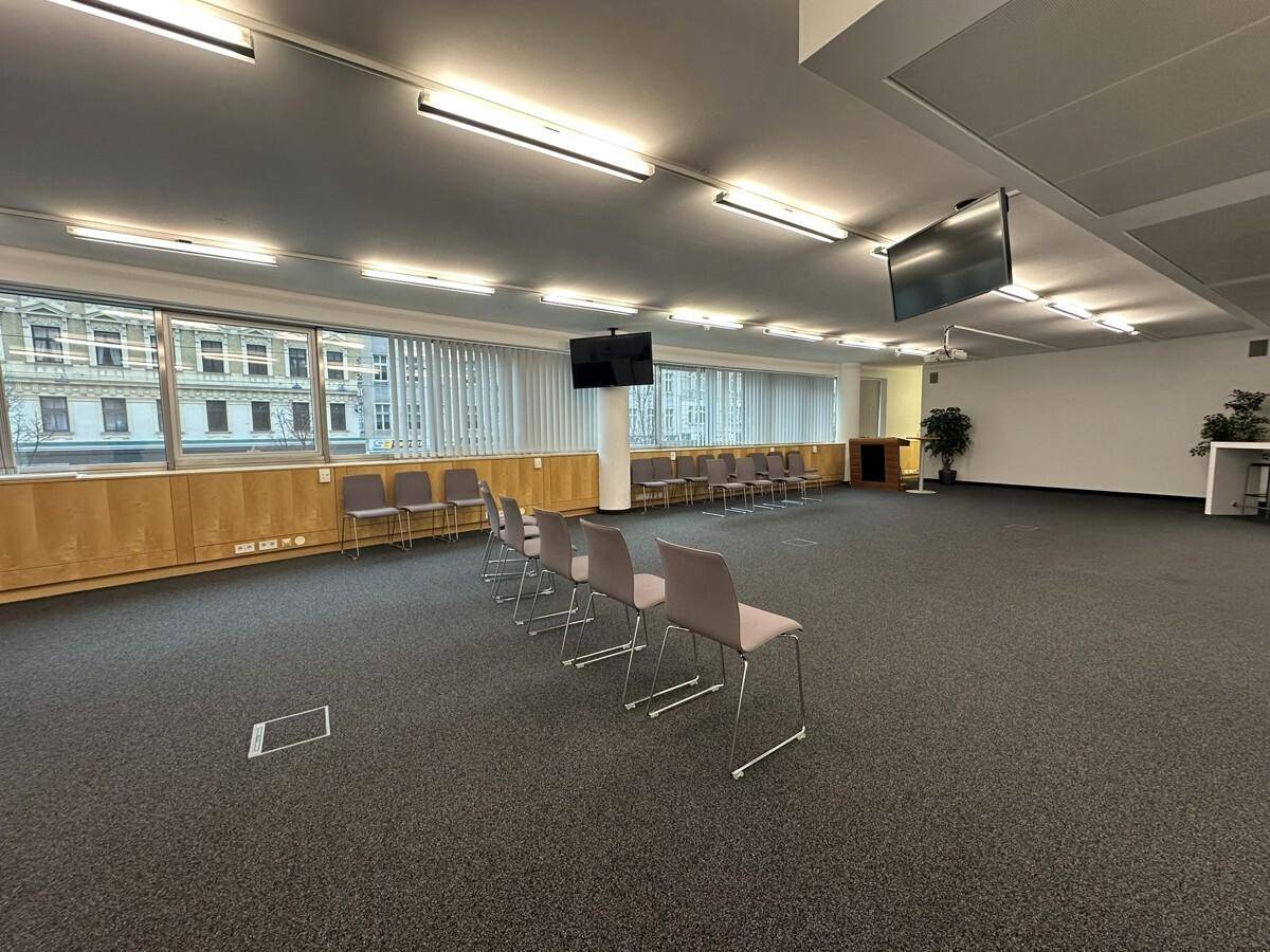 Großer Konferenzraum mit Reihen von Stühlen, großen Fenstern und an der Wand montierten Bildschirmen für Präsentationen.