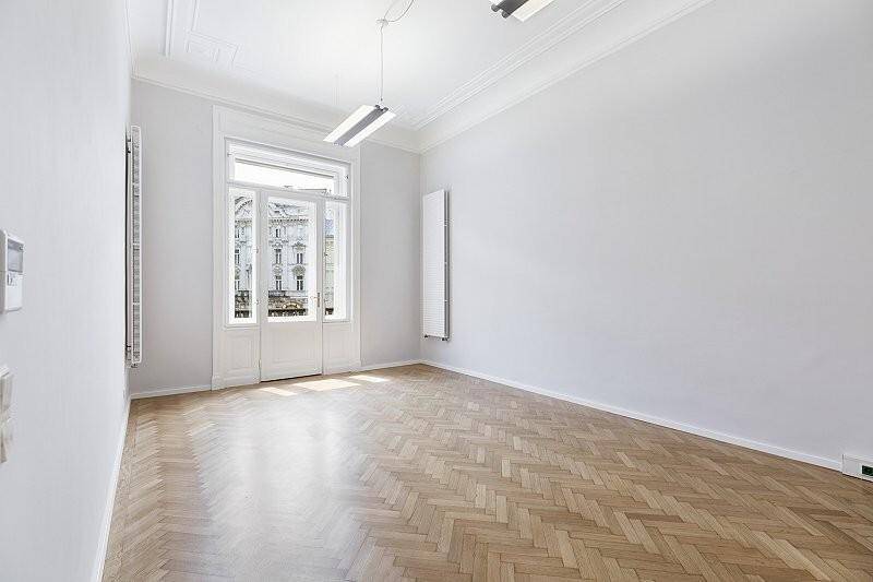 Geräumiger Raum mit Fischgrätparkett, hohen Decken und Zugang zu einem Balkon im Obergeschoss.