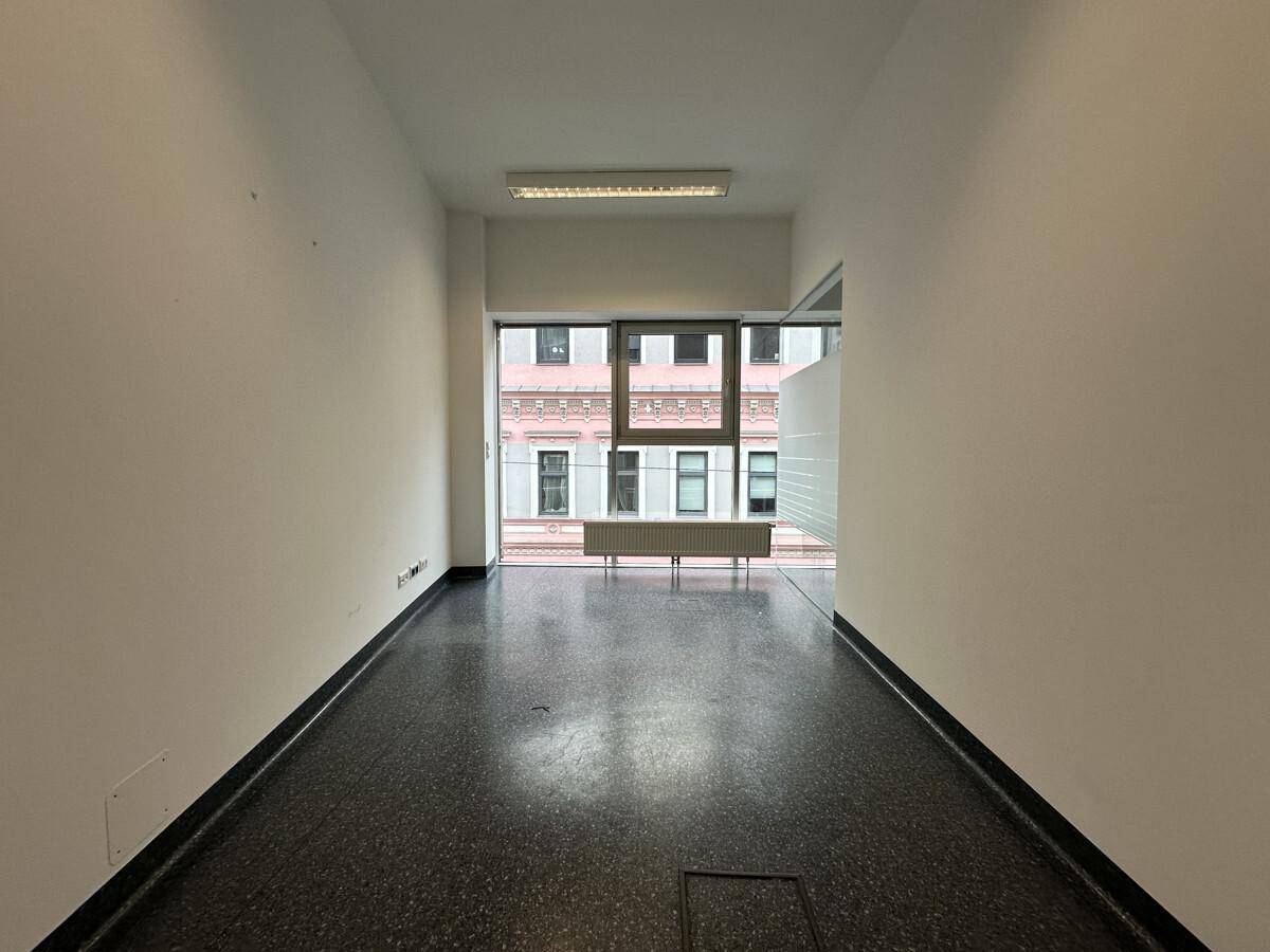 Schmaler Büroraum mit Fenster, Blick auf die gegenüberliegenden Gebäude und dunklem Fliesenboden.
