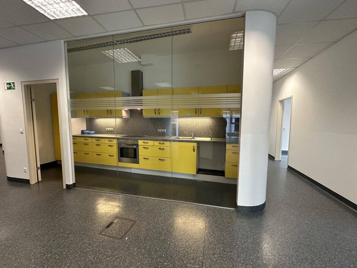 Moderne Einbauküche mit gelben Schränken, Edelstahlgeräten und Glaswand in einem Bürobereich.