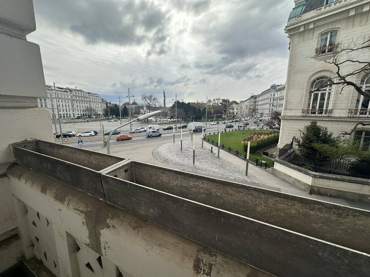 Blick von einem Balkon auf eine belebte Stadtstraße mit Verkehr und historischen Gebäuden unter bewölktem Himmel.