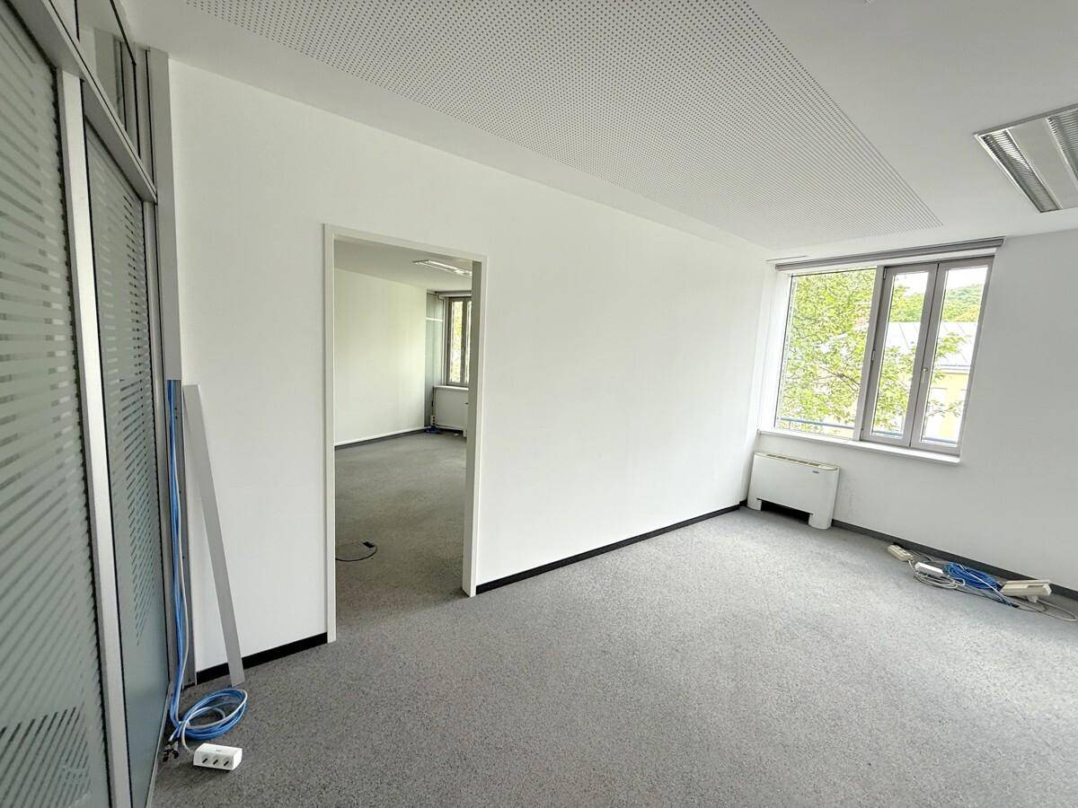 Büroraum mit Teppichboden, weißen Wänden und einer Glastrennwand, die zu einem weiteren Raum führt.