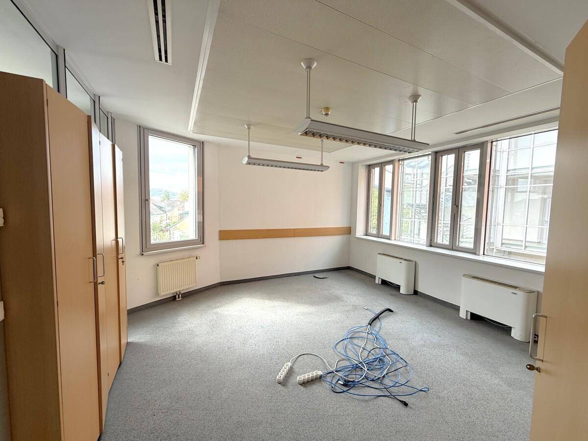 Helles Büro mit grauem Teppichboden, weißen Wänden, Holzschränken und Fenstern mit Ausblick.