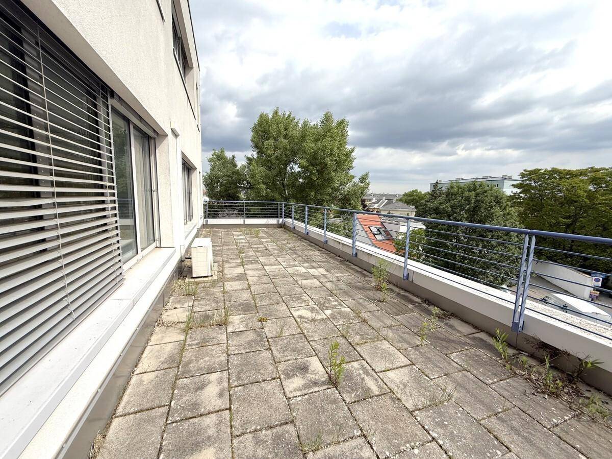 Große Außenterrasse mit Betonplatten und Metallgeländer, bietet einen Blick auf Bäume und Gebäude.