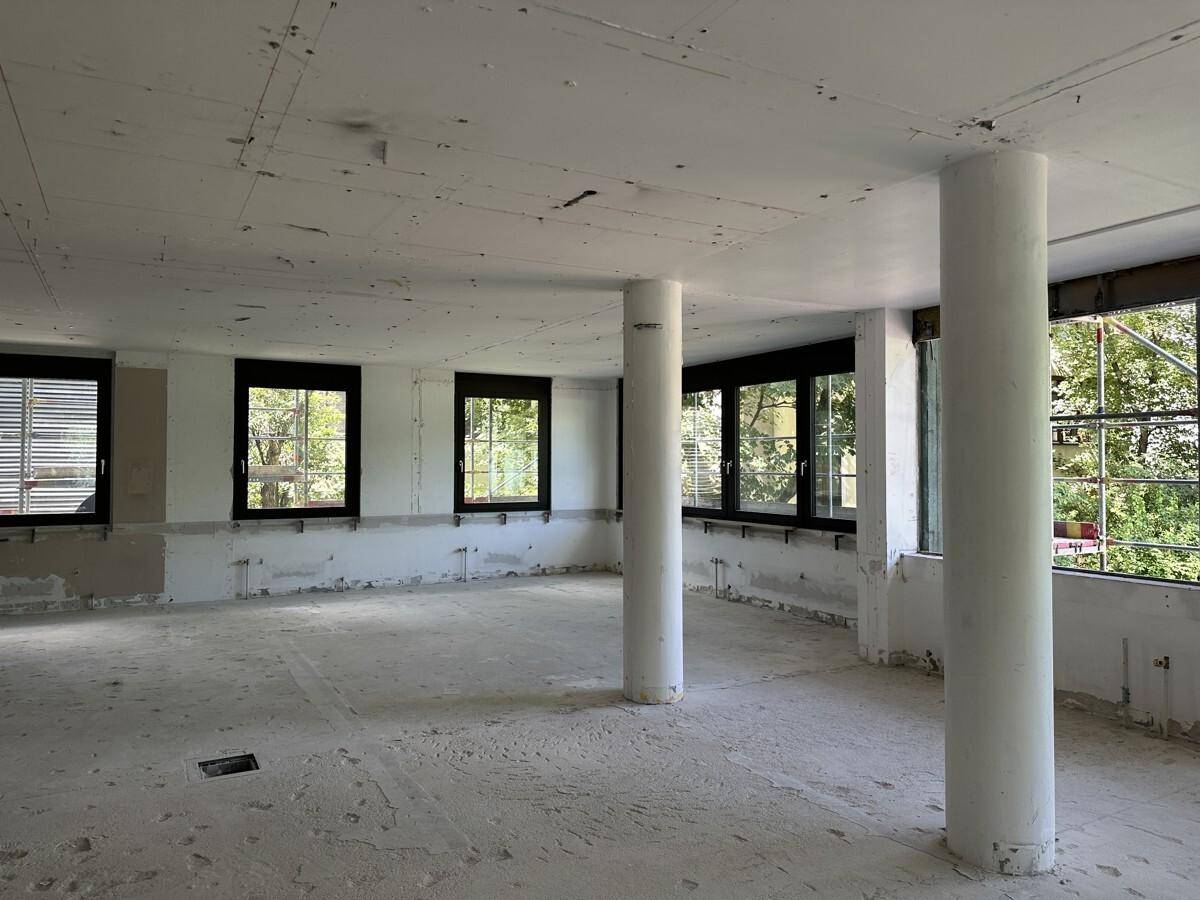 Ein großer, leerer Raum in Renovierung mit Säulen und vielen Fenstern mit Blick ins Grüne.