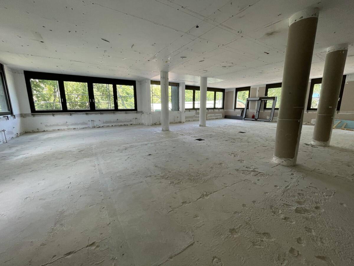 Geräumiger Büroraum im Rohbau mit mehreren Säulen und großen Fenstern, die viel Licht hereinlassen.