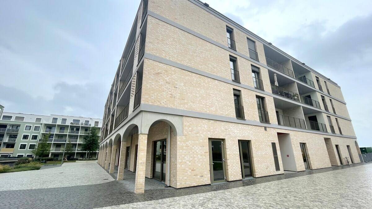 Seitenansicht eines Neubaus mit heller Ziegelfassade, Balkonen und großen Fenstern im Erdgeschoss.