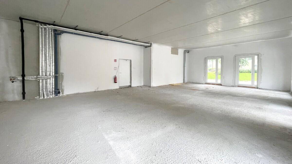 Unausgebauter Gewerberaum mit sichtbaren Installationen an der Wand und großen Fenstern.