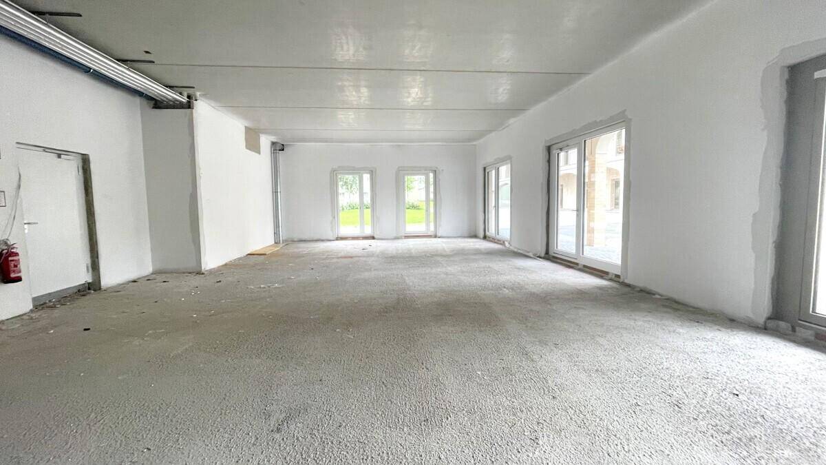 Geräumiger Gewerberaum im Rohbau mit Betonboden und mehreren Fenstern, bereit für den Ausbau.