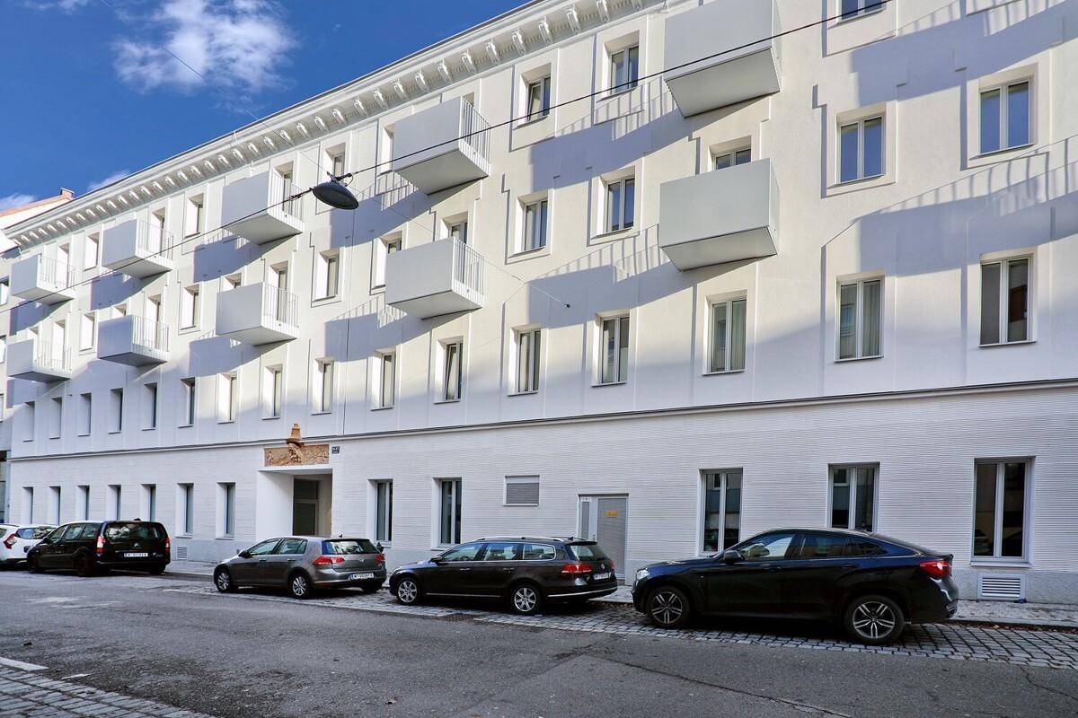 Weiße Gebäudefassade mit vielen kleinen Balkonen und parkenden Autos auf der Straße davor.