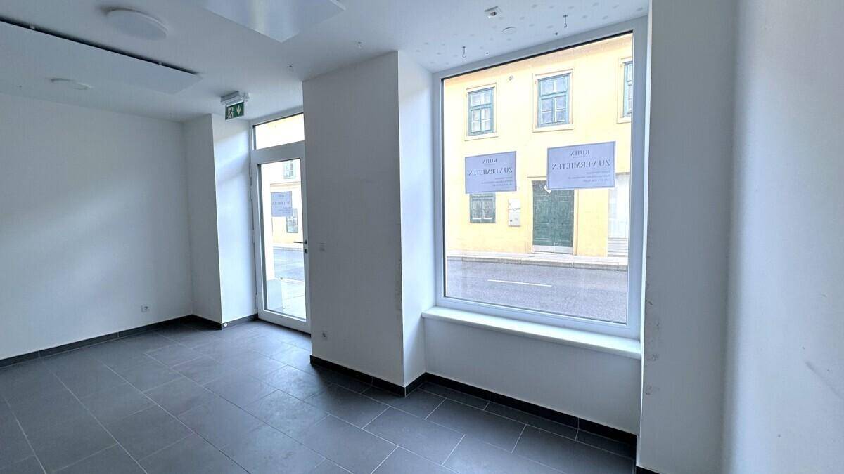 Heller, leerer Geschäftsraum im Erdgeschoss mit großen Fenstern zur Straße und Fliesenboden.