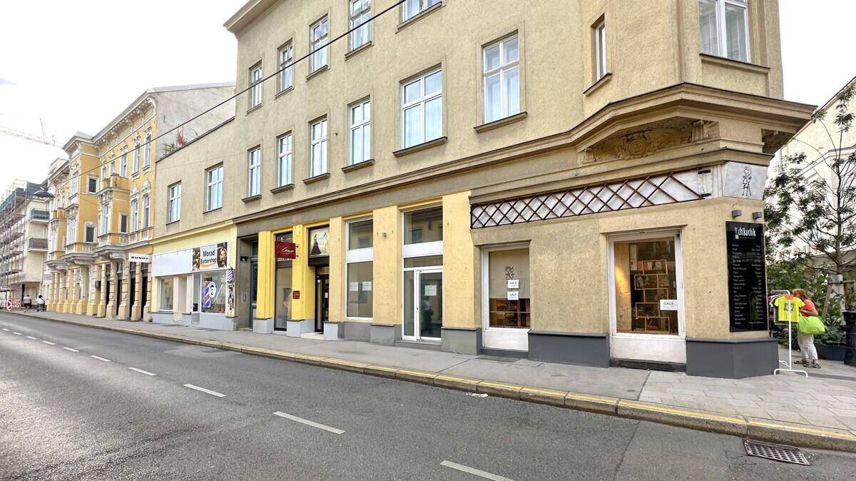 Ansicht der Gebäudefassade mit Ladenlokalen im Erdgeschoss und darüberliegenden Etagen, entlang einer städtischen Straße.