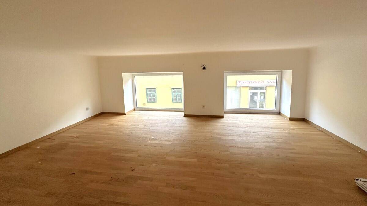 Großer, heller Raum mit Holzboden und zwei breiten Fenstern, die viel Tageslicht hereinlassen.