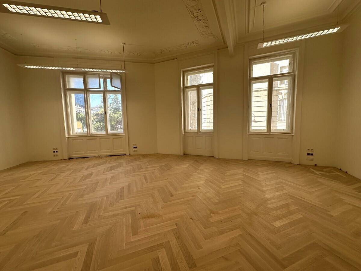 Geräumiges Büro mit Fischgrätparkett, hohen Decken und großen Fenstern, die viel Tageslicht hereinlassen.