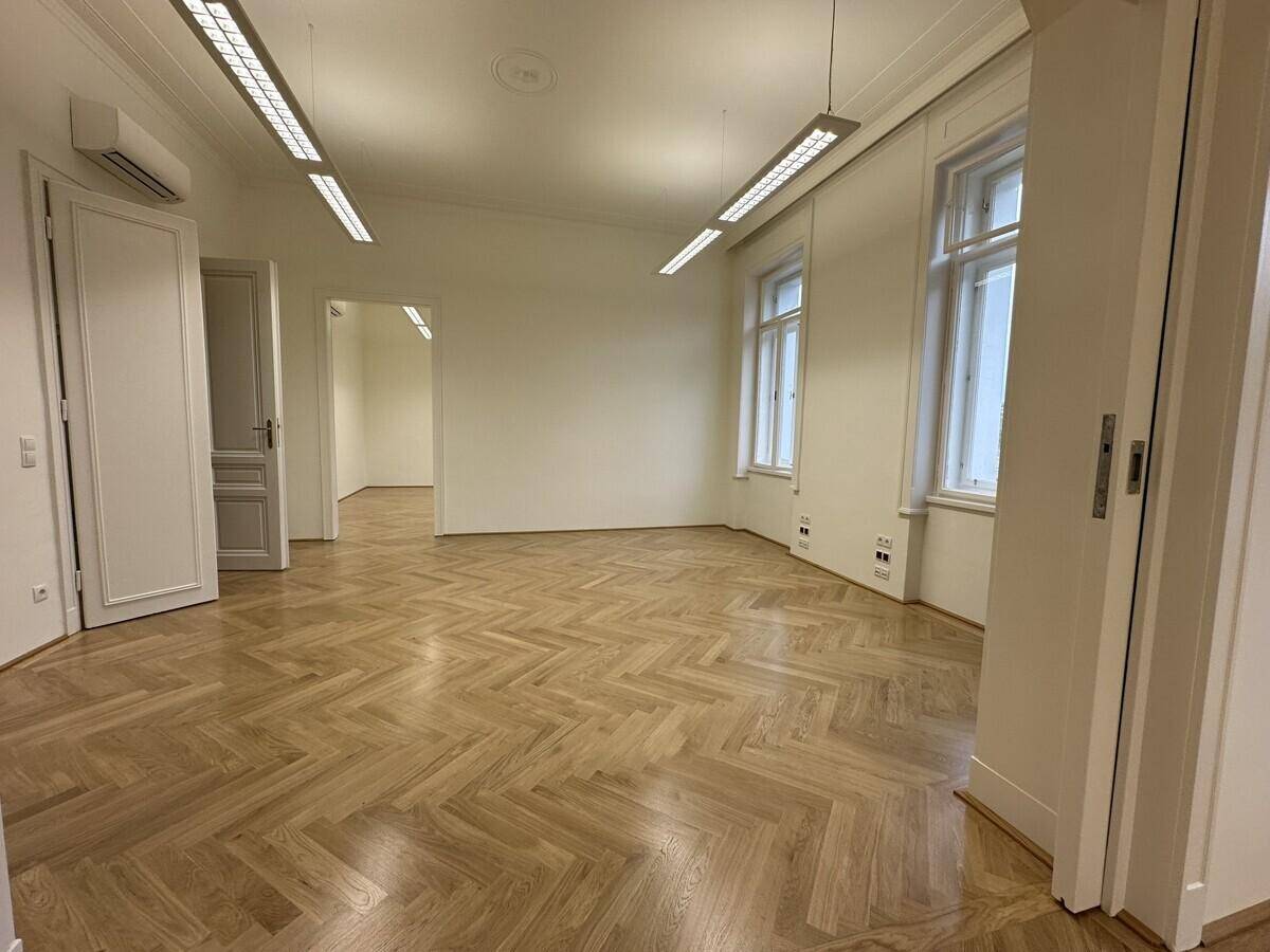 Geräumiges Büro mit Fischgrätparkett, Klimaanlage und mehreren Fenstern, die den Raum erhellen.