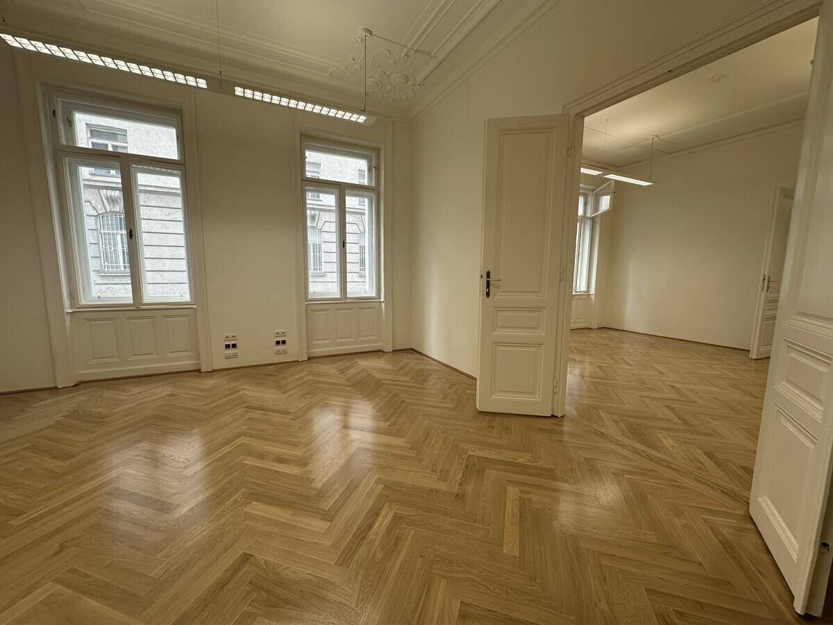 Helles Büro mit Fischgrätparkett, hohen Fenstern und einer offenen Tür, die den Blick in einen weiteren Raum freigibt.