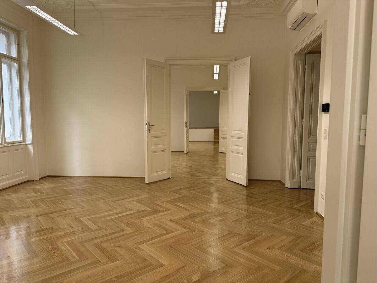 Geräumiges Büro mit Fischgrätparkett, Klimaanlage und mehreren Fenstern, die den Raum erhellen.