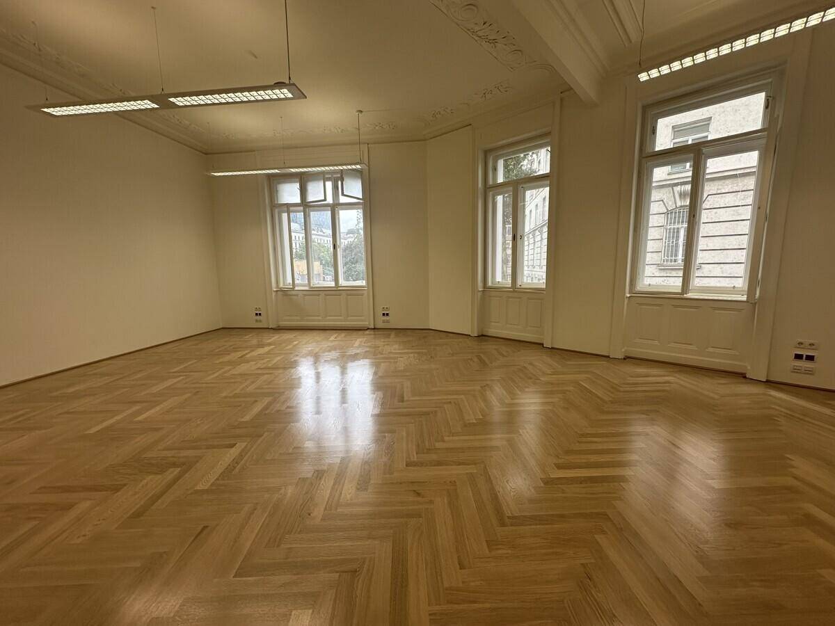 Geräumiges Büro mit Fischgrätparkett, hohen Decken und großen Fenstern, die viel Tageslicht hereinlassen.