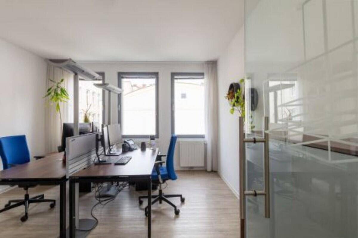 Heller Büroraum mit zwei Arbeitsplätzen, großen Fenstern und modernen Möbeln.