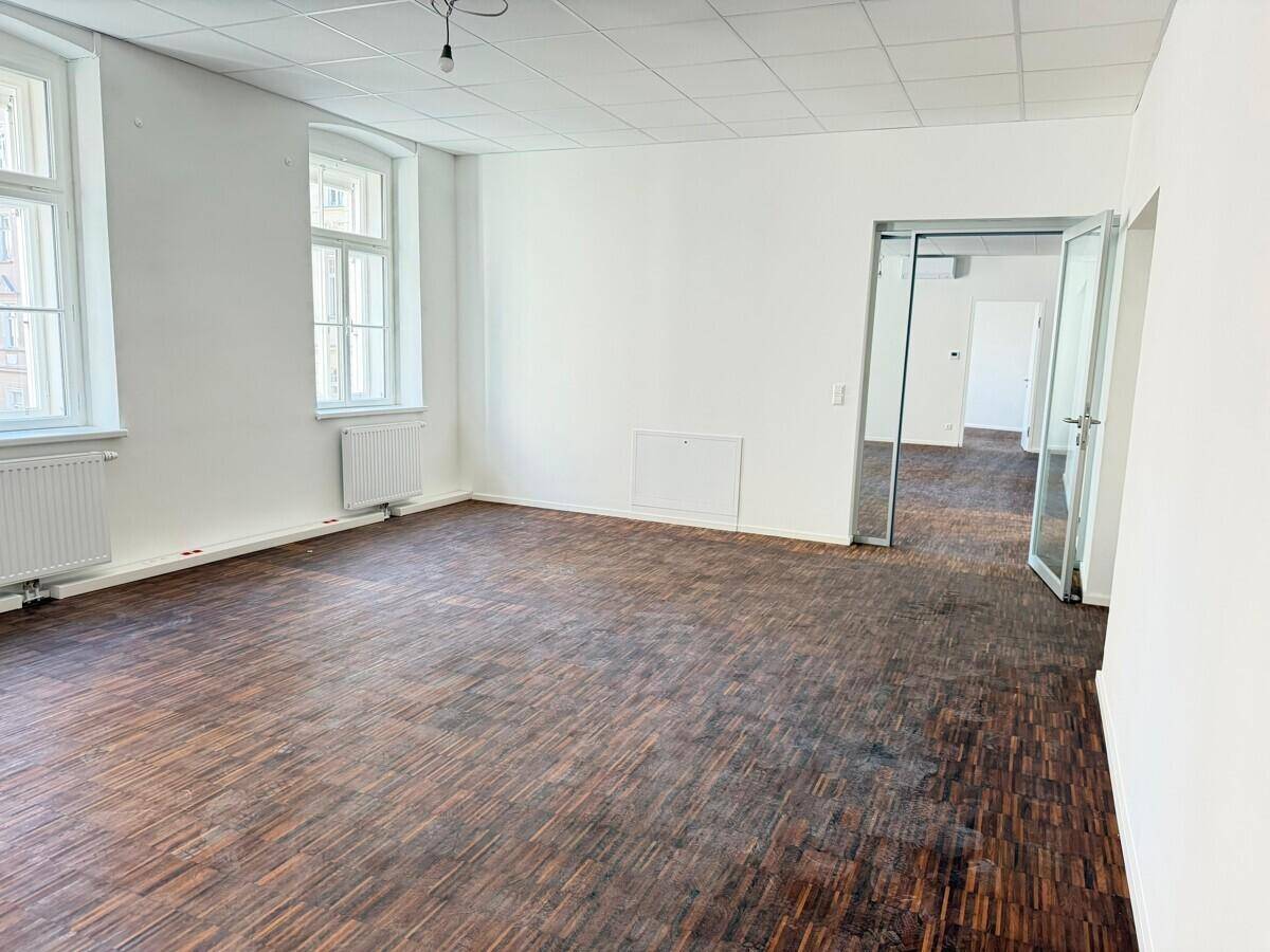 Geräumiger Büroraum mit dunklem Holzparkett, weißen Wänden und großen Fenstern, bereit zur Einrichtung.