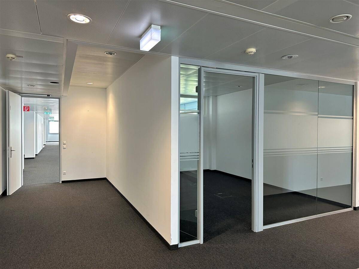 Heller Büroflur mit Teppichboden und modernen Glaswänden, die separate Büroräume abgrenzen.