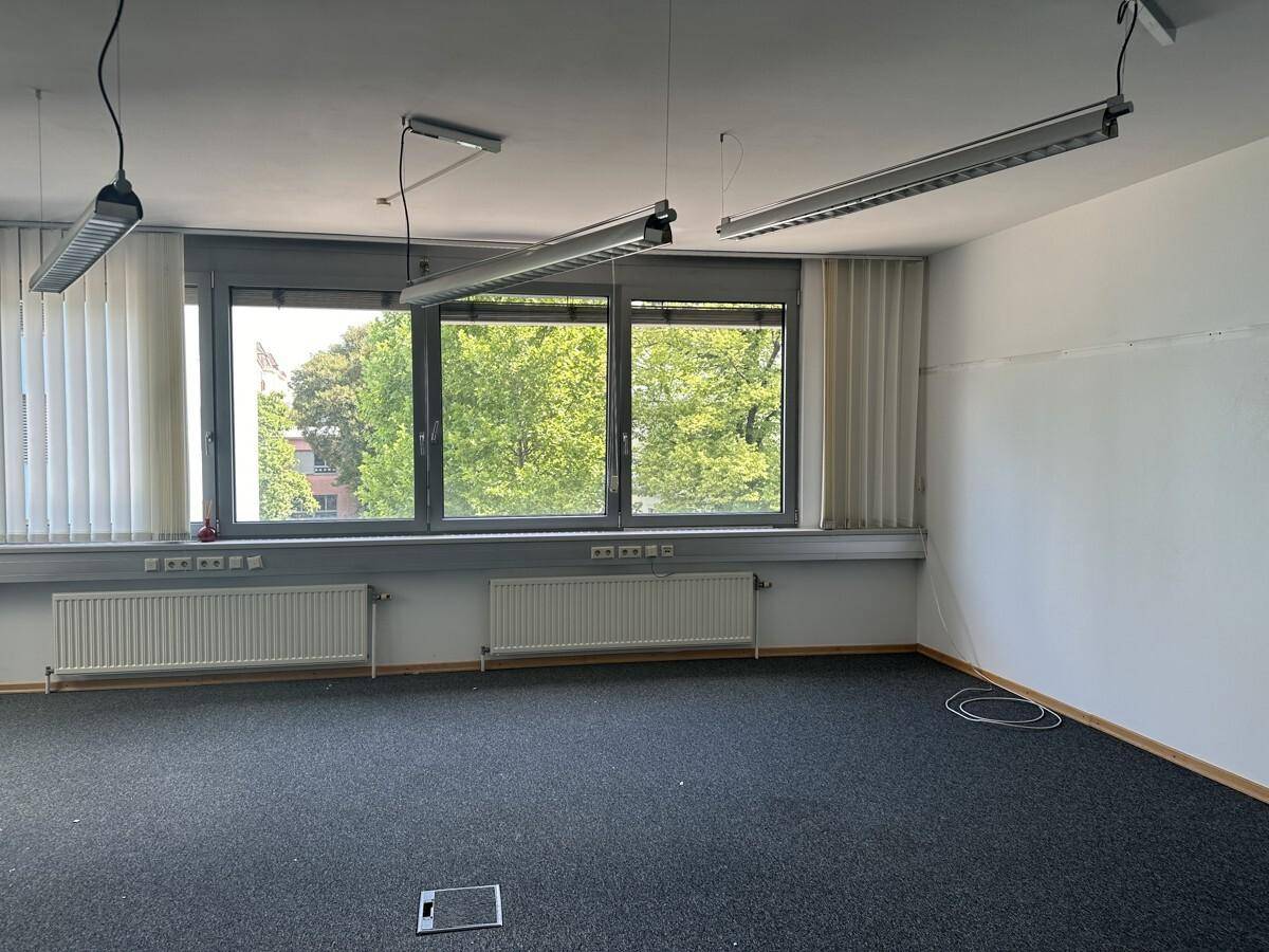 Leeres Büro mit dunklem Teppichboden, großen Fenstern mit Jalousien und Heizkörpern unter den Fenstern.