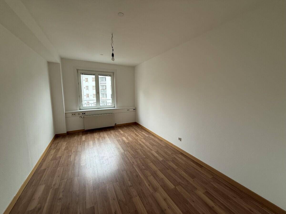 Heller, leerer Raum mit Laminatboden und einem großen Fenster, das viel Tageslicht hereinlässt.