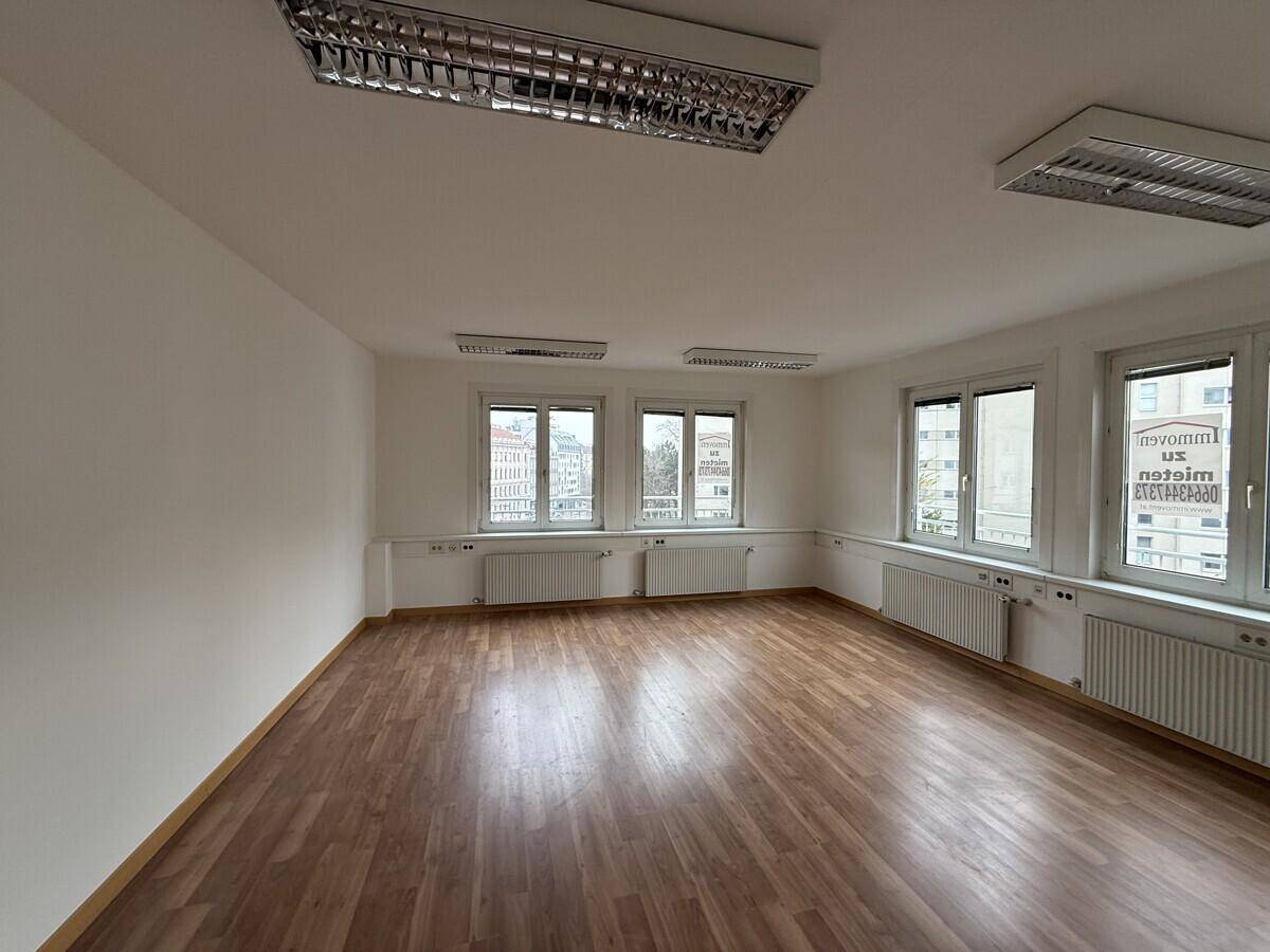 Geräumiger, leerer Raum mit Laminatboden, weißen Wänden und mehreren Fenstern, ideal für Büronutzung.