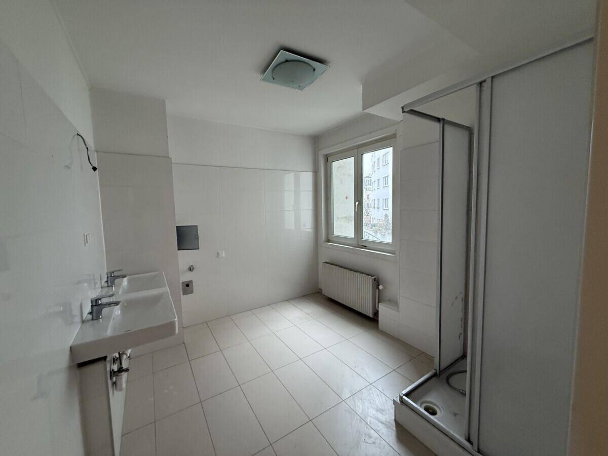 Helles Badezimmer mit weißen Fliesen, zwei Waschbecken und einer Duschkabine sowie einem Fenster.