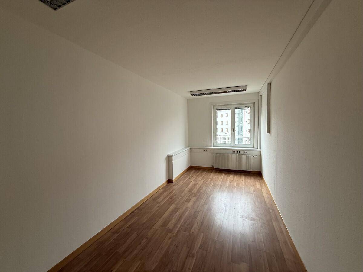Schmaler Büroraum mit Laminatboden, weiß gestrichenen Wänden und einem Fenster mit Stadtblick.