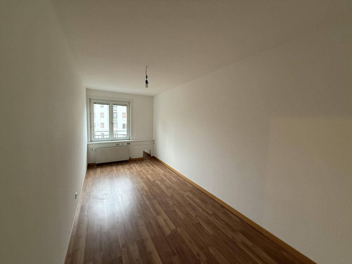 Leerer Raum mit Laminatboden und einem Fenster, das den Blick auf die umliegenden Gebäude freigibt.