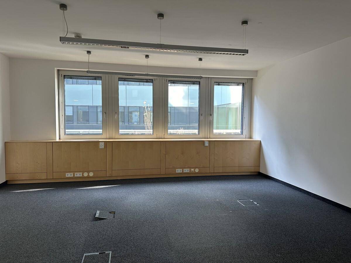 Leeres Büro mit Teppichboden, großen Fenstern und integrierten Holzschränken unter den Fenstern.