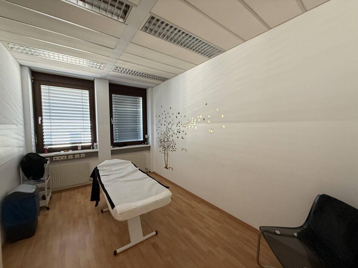 Behandlungsraum mit einer Liege, Holzboden und zwei Fenstern, dekoriert mit einem Baum-Wandtattoo.