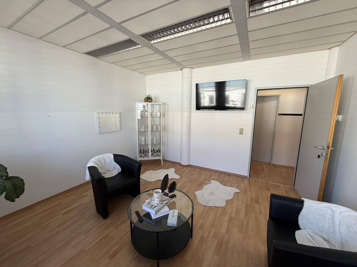 Heller Empfangsbereich oder Wartezimmer mit zwei schwarzen Sesseln, einem Couchtisch und einem Wandfernseher.