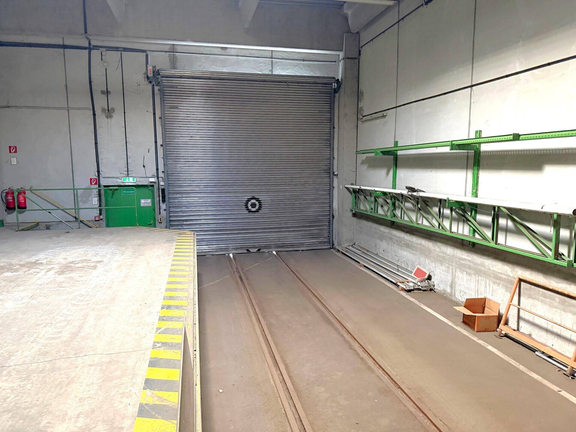 Große Laderampe mit Rolltor und Schienen im Boden, umgeben von industriellen Regalsystemen.