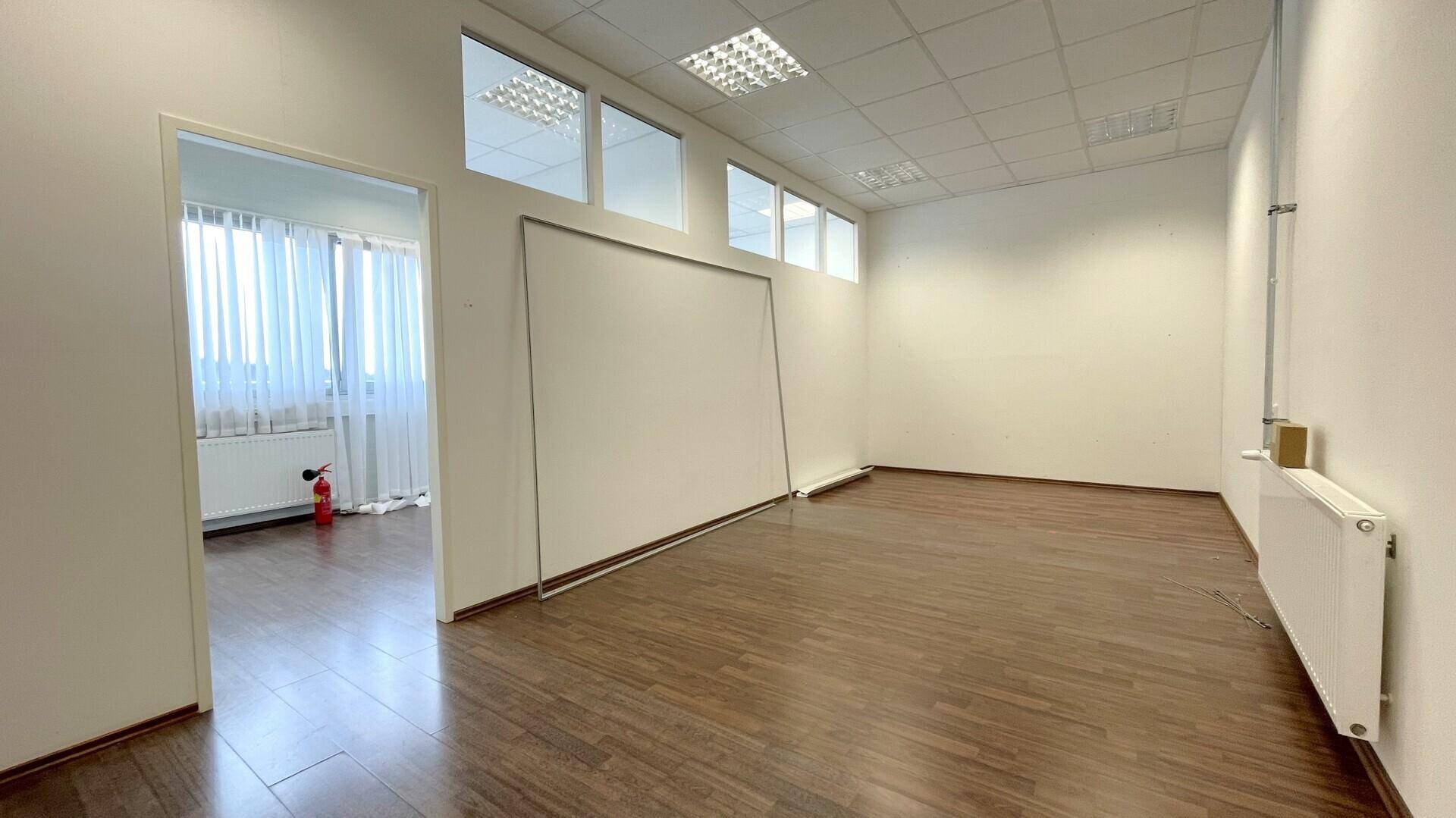 Geräumiger, leerer Büroraum mit Holzboden, weißen Wänden und Deckenplatten mit integrierter Beleuchtung.