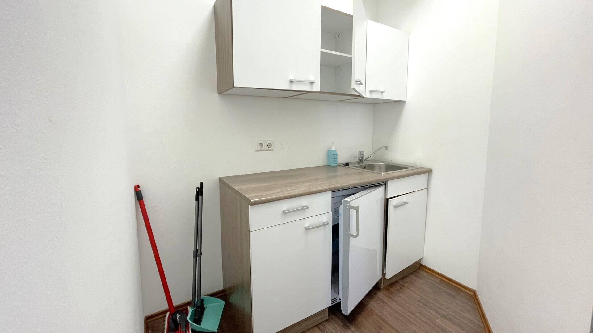 Kleine Küchenzeile mit weißen Schränken, Spüle, Kühlschrank und Holz-Arbeitsplatte.