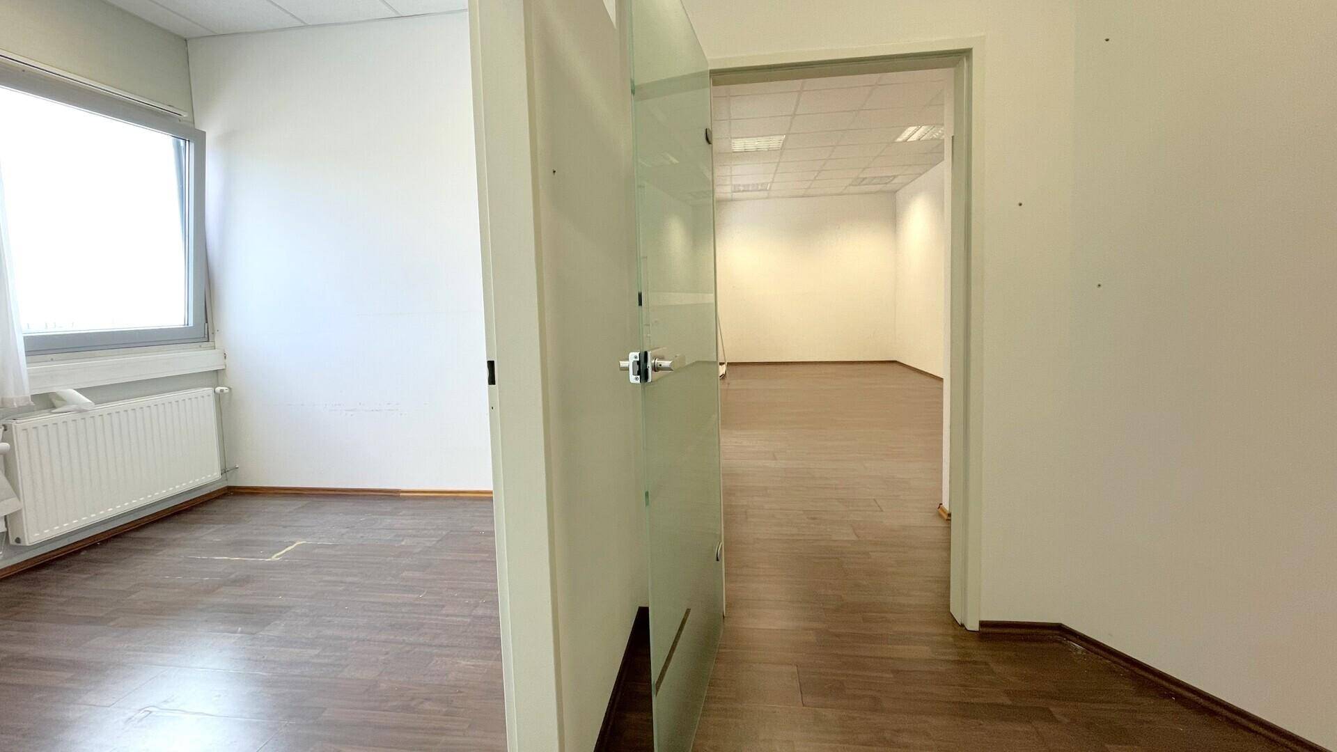 Blick aus einem Flur in einen leeren Büroraum mit großen Fenstern und Holzboden.