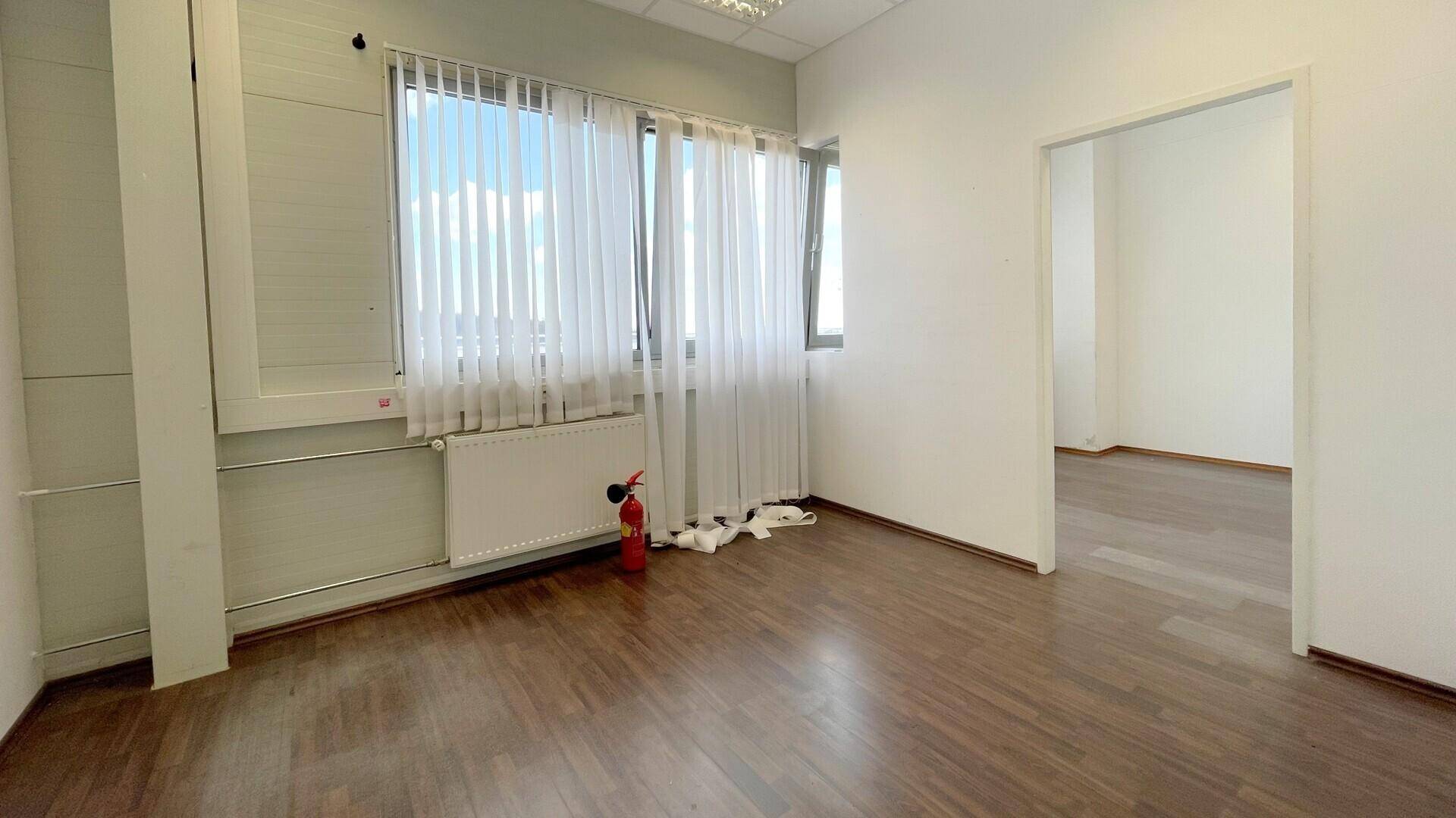 Leerer Büroraum mit Holzboden, großen Fenstern, Heizkörper und einem Feuerlöscher an der Wand.