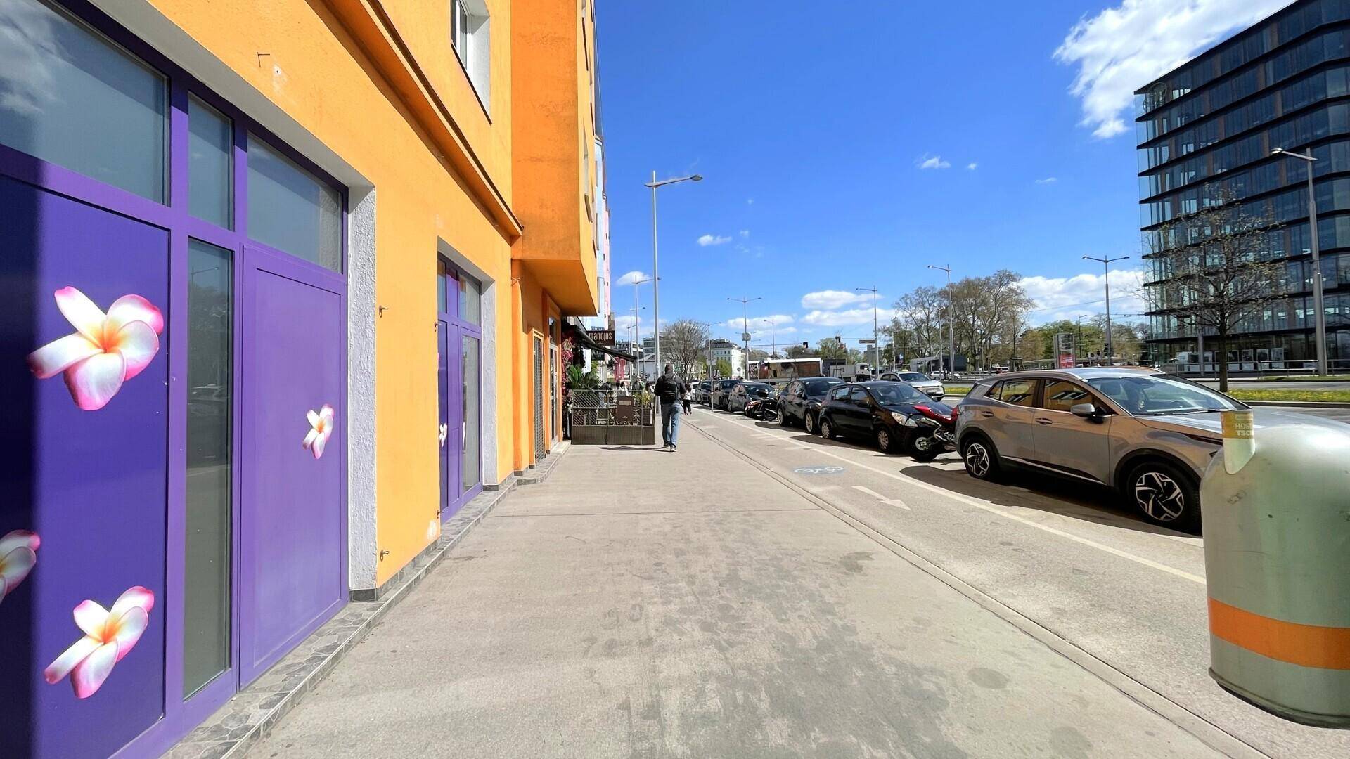 Bürgersteig vor einem farbenfrohen Gebäude mit Geschäften und parkenden Autos entlang der Straße.