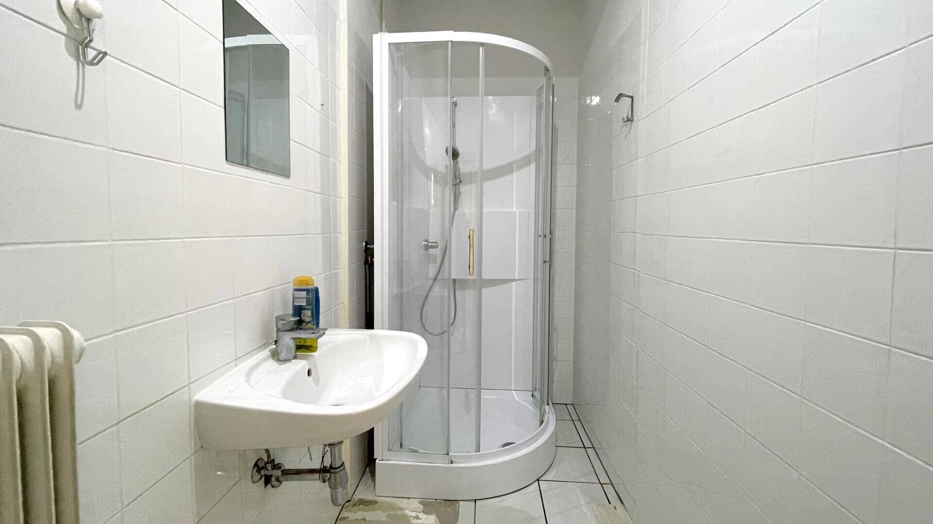 Helles Badezimmer mit weißen Fliesen, einer Duschkabine und einem Waschbecken an der Wand.