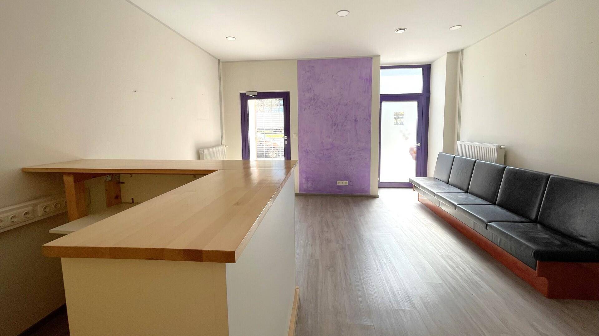 Heller Empfangsbereich mit einer Holztheke, Sitzgelegenheiten und großen Fenstern zur Straße.