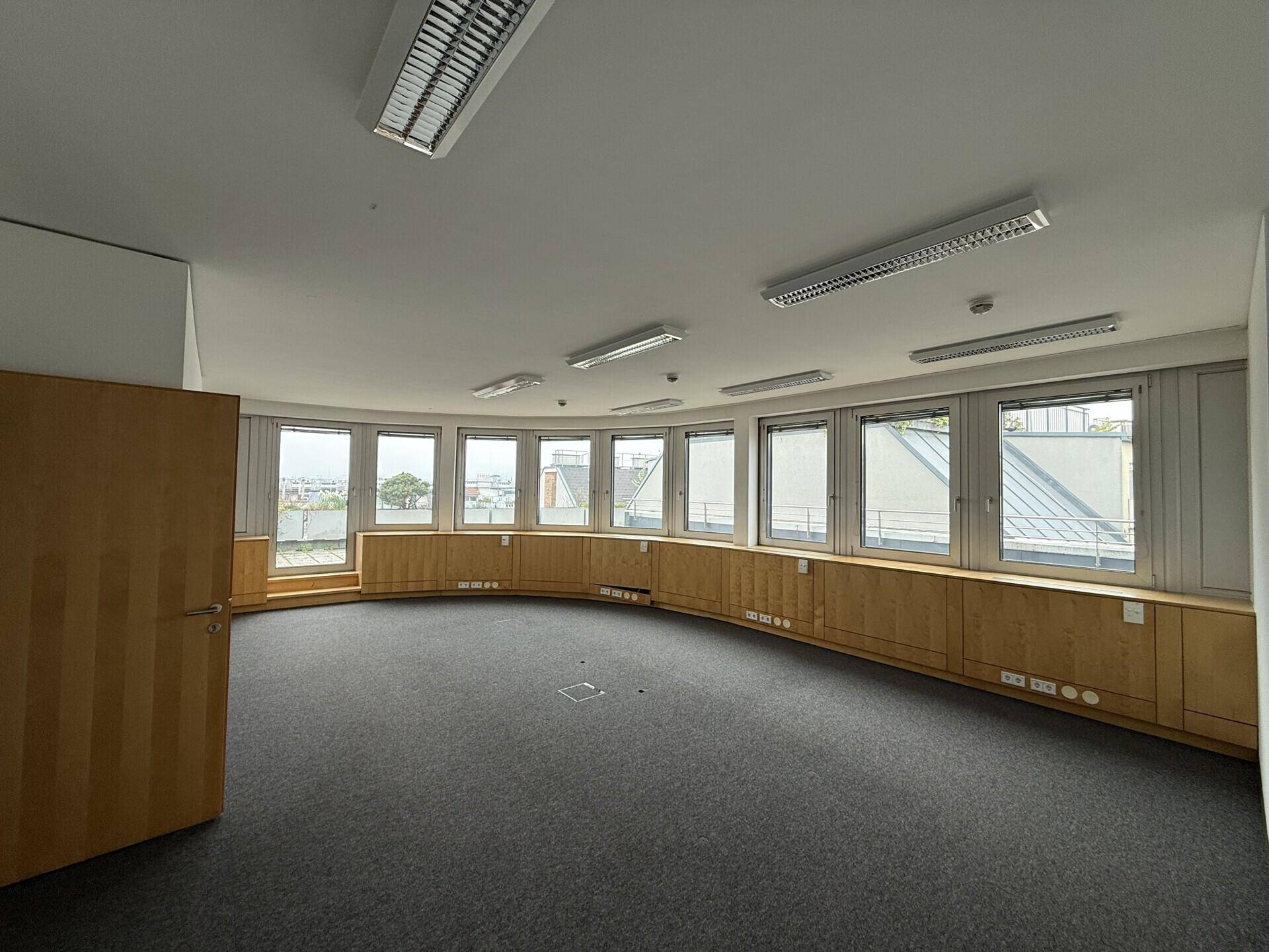 Heller Büroraum mit Teppichboden, gebogener Fensterfront und Blick auf die Dächer der Stadt.