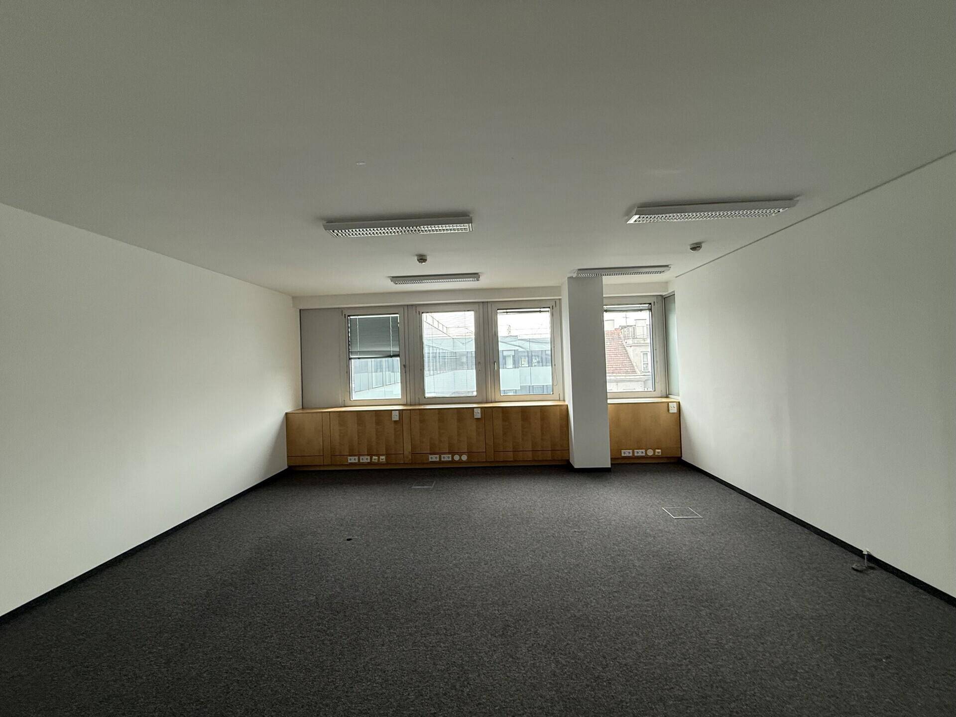 Leerer Büroraum mit Teppichboden, Holzvertäfelung unter den Fenstern und neutralen Wänden.