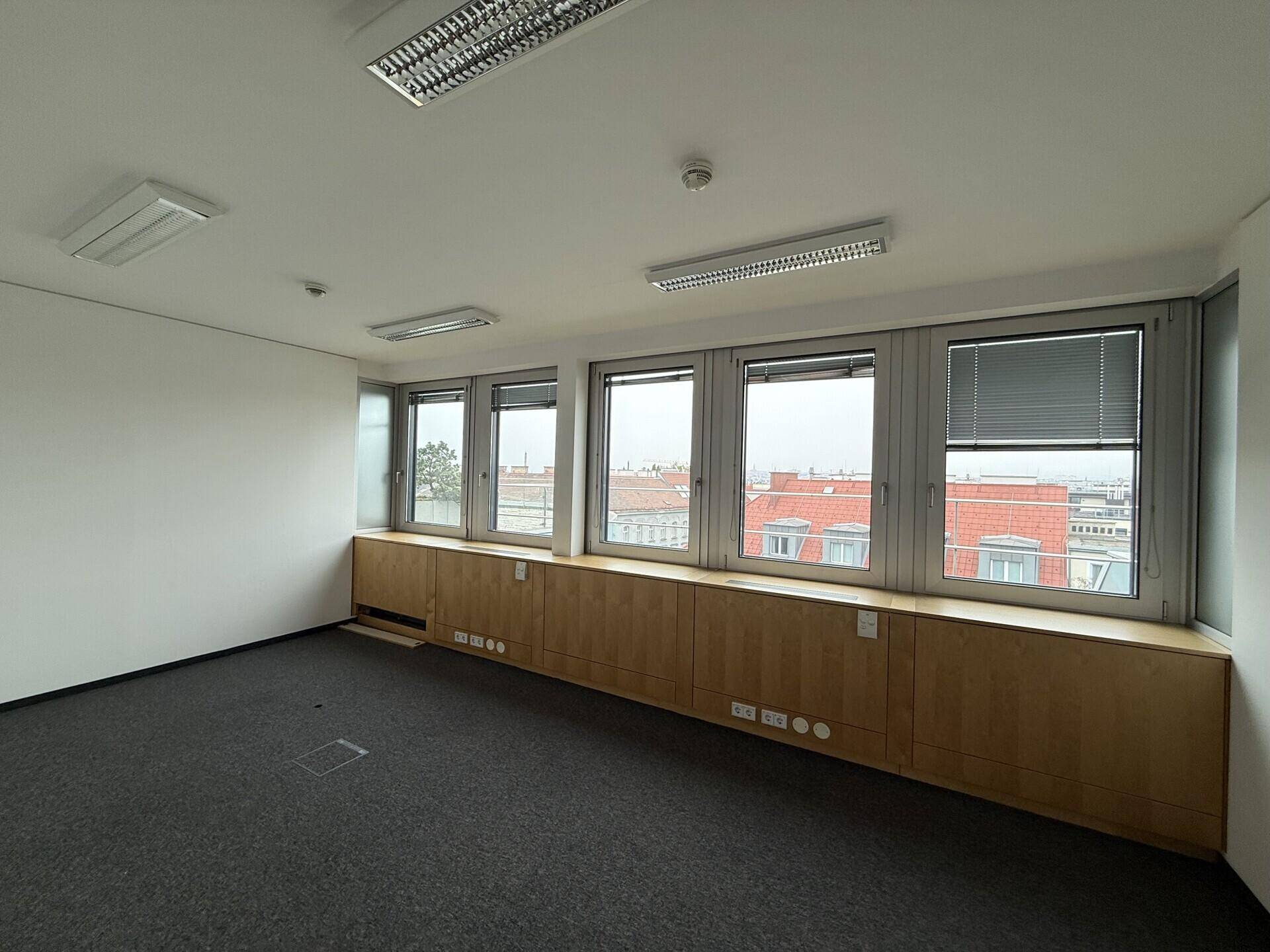 Geräumiger Büroraum mit Teppichboden, breiter Fensterfront und Holzverkleidung unter den Fenstern.