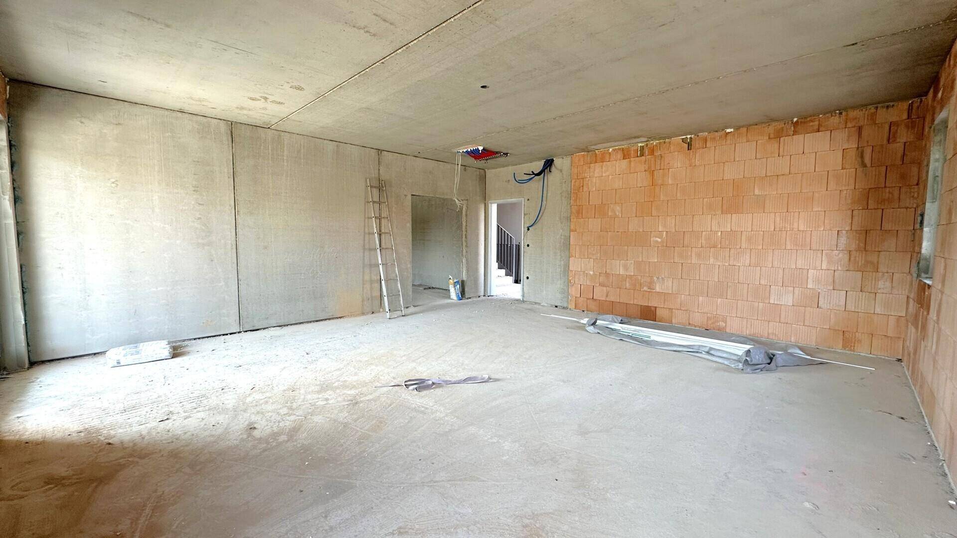 Großer Rohbau-Raum mit Ziegel- und Betonwänden, Betonboden und Blick auf einen Treppenaufgang.