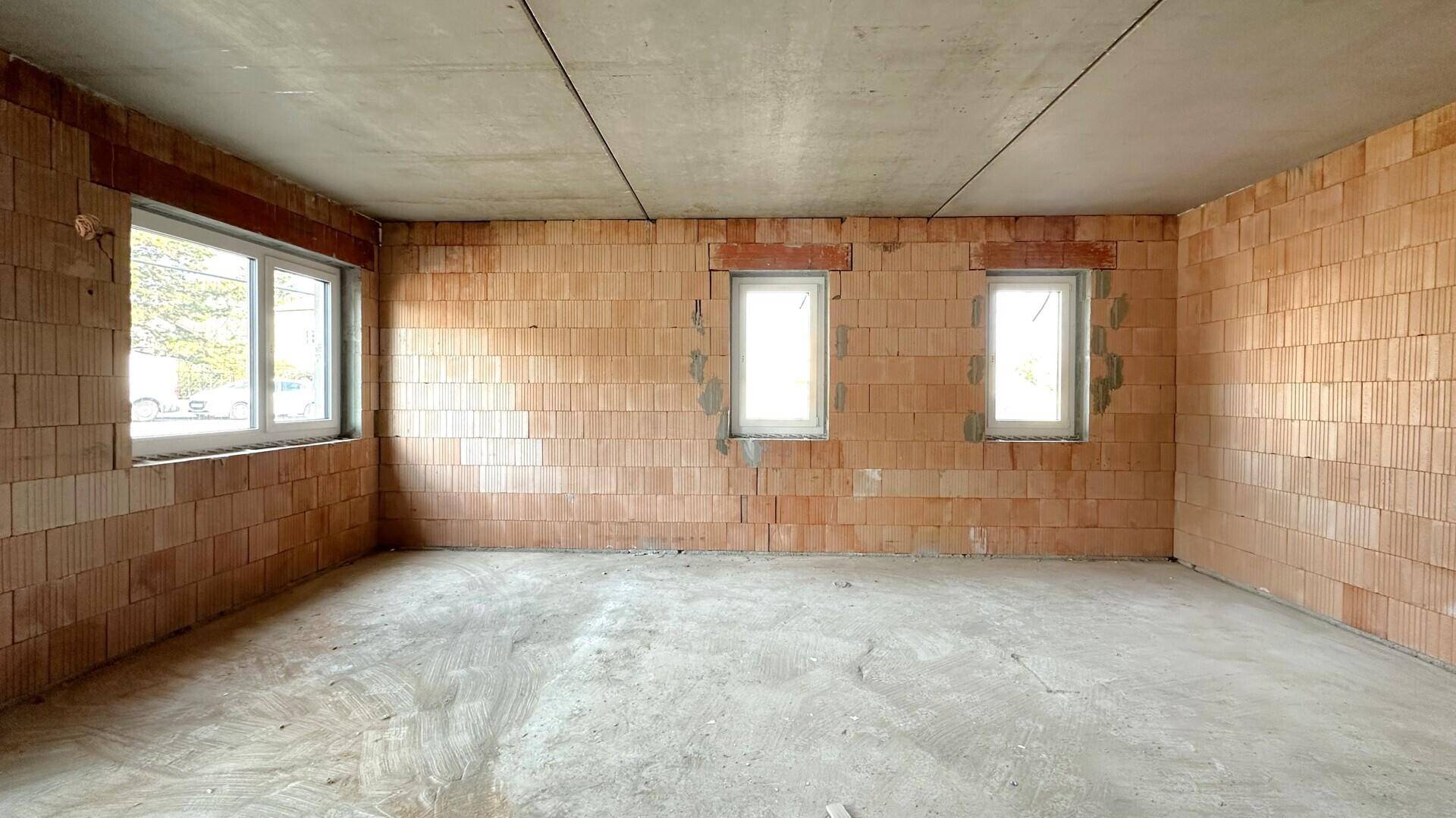 Unfertiger Raum im Rohbauzustand mit Ziegelwänden, Betonboden und mehreren Fenstern, die Licht hereinlassen.